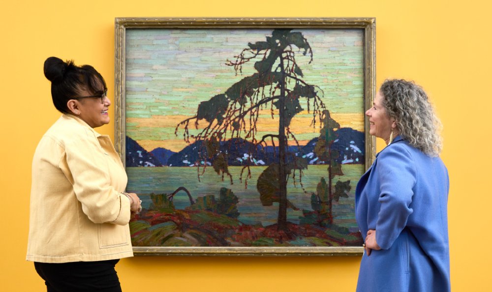 Medium shot of two individuals looking at Tom Thomson's The Jack Pine, hung on a yellow wall.