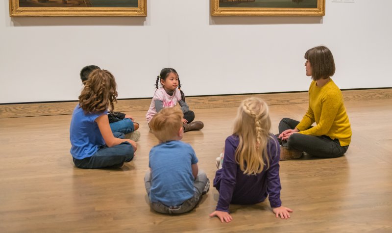 Students sitting on the ground in front of artwork.